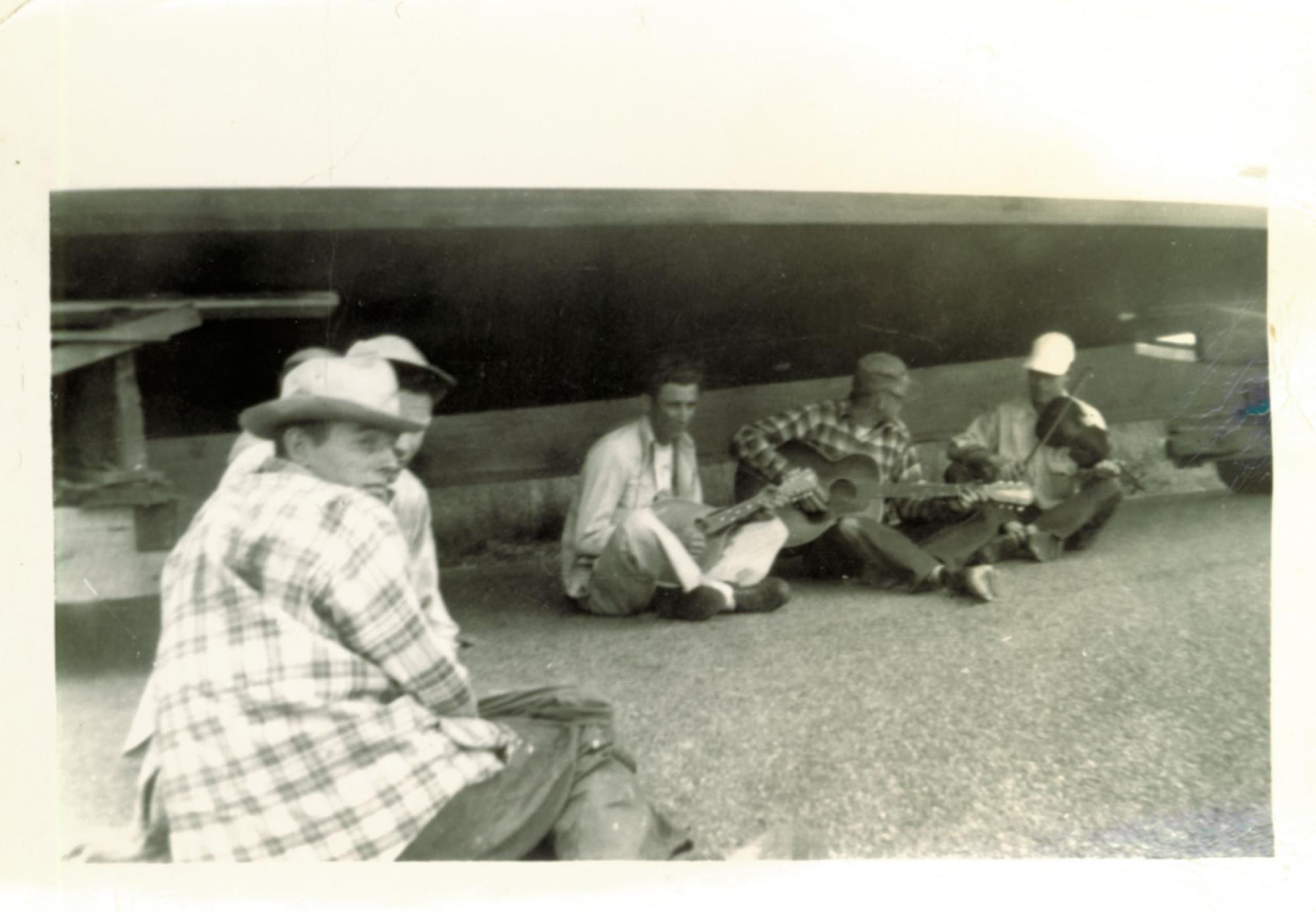Bronza boatyard crew members Tom Dean, O'Neal Dean, and Ralph Ruark play mandolin, guitar, and fiddle while waiting for tide to come up to launch. Unidentified men in the foreground may be Bronza's clients. Undated, probably 1954-56.