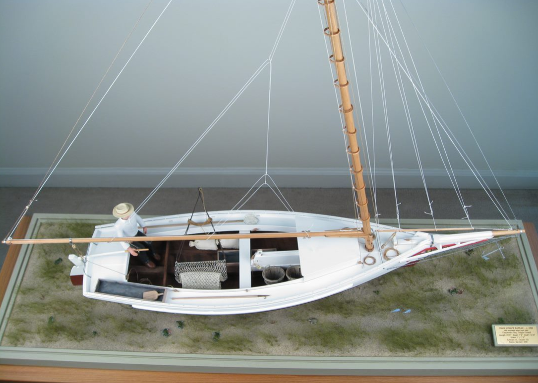 On port washboard – crab dip-net. Under that are two sandbags, each about 40-50pounds, used to balance the boat against the wind. An empty box and two bushel-baskets are ready to receive caught crabs. On the starboard washboard is a “dump box” – the scrape net contents are “dumped” into this box for culling, sorting out the keeper crabs, and returning the rest overboard. In the dump box is a carved bailing scoop, and forward of that is a boat hook. (The hook is carved from a bronze boat screw, over 100 yrs old.) The scrape occupies the rest of boat.